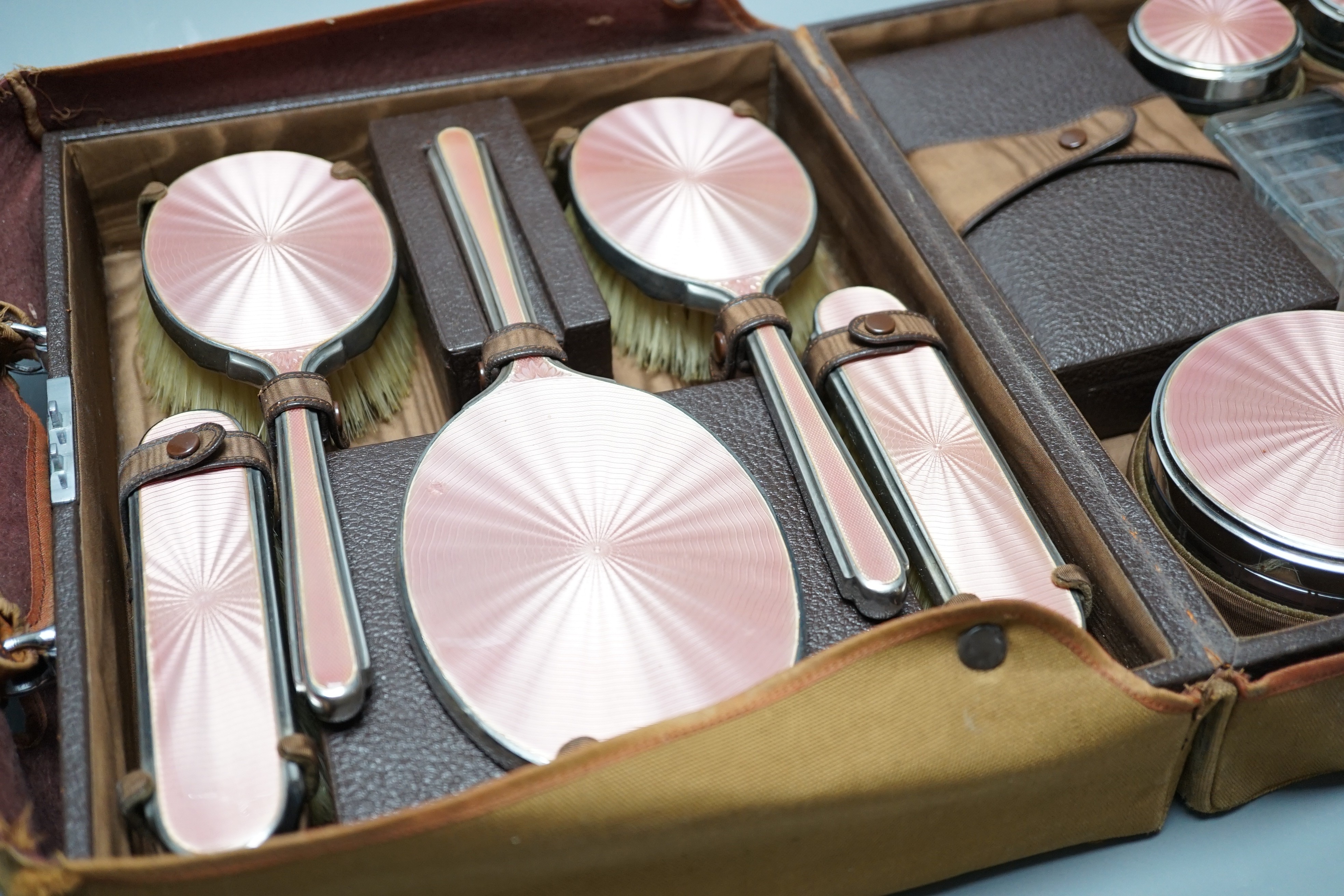 A George V travelling toilet case, containing eleven silver and pink enamel mounted glass bottles jars, mirror and brushes, Mappin & Webb, 1936, in brown leather case, case width 31cm.